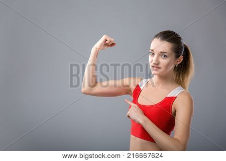 Beautiful stylish young woman is pointing on her muscles and smiling, on gray background. space for text