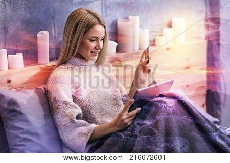 Modern technology. Cheerful attractive pleasant woman lying under the blanket and holding a tablet while doing online shopping