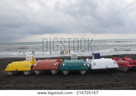 Colorful series of pedal boat parked on the beach overcast clouds waves. Blurred photo of people on beach