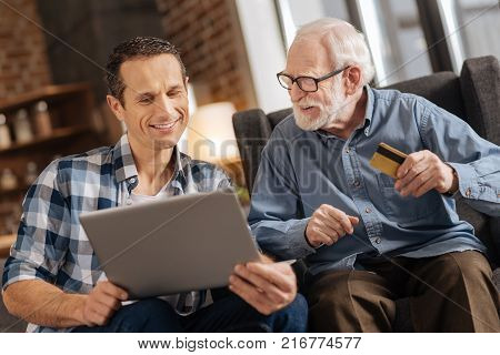 Fast and easy. Pleasant young man holding a laptop and showing an online shop webpage to his elderly father while the elderly man holding a bank card, ready to pay for purchases