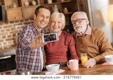 Best moments. Cheerful family consisting of an adult son and elderly parents taking selfies while having breakfast in the kitchen