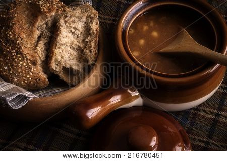 Food, a soup broth  and seeded Roll winter warming lunch, showing a rustic food scene with wooden bread dish and earthenware crockery soup dish with lid and wooden spoon.