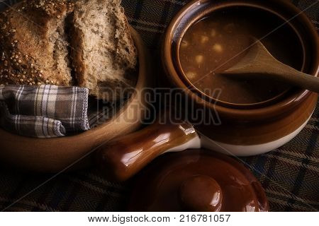 Food, a soup broth  and seeded Roll winter warming lunch, showing a rustic food scene with wooden bread dish and earthenware crockery soup dish with lid and wooden spoon.