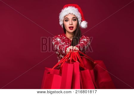 Young pretty woman or girl with long beautiful hair in Santa hat and New Year's dress holding red package on red studio background. Christmas sale. Emotional brunette. Copy space