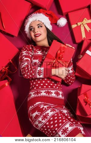 Funny young woman in a New Year's dress lying on a mountain of gifts. Christmas sale. Emotional brunette in Santa hat