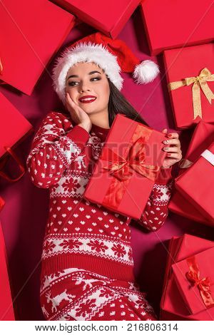 Funny young woman in a New Year's dress lying on a mountain of gifts. Christmas sale. Emotional brunette in Santa hat