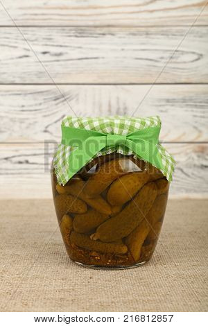 Close up of one glass jar of pickled small gherkin cucumbers checkered linen textile fabric lid decoration and green ribbon on canvas over white wooden background low angle side view