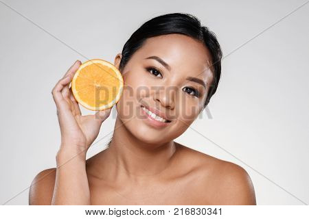 Attractive half-naked asian woman amiling while holding orange slices near her face isolated over grey