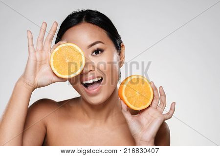 Attractive half-naked asian woman amiling while holding orange slices near her face isolated over grey