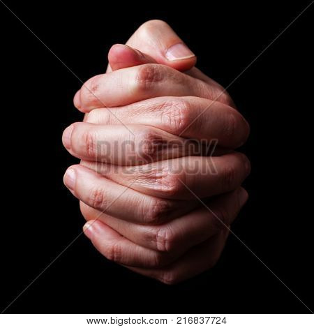 Low key, close up of hands of a faithful mature man praying, hands folded, interlaced fingers in worship to god. Isolated black background. Concept for religion, faith, prayer and spirituality.