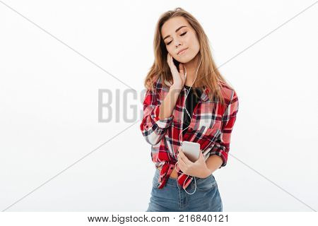 Portrait of a satisfied pretty girl in plaid shirt holding mobile phone and listening to music with earphones isolated over white background