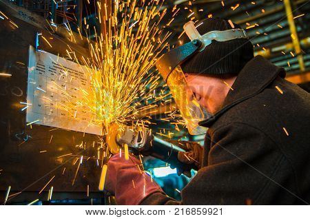 A man in working form and a face mask cuts a metal saw with a Bulgarian on the background of a metallurgical industrial plant.