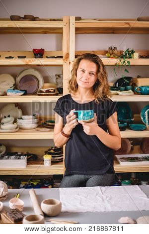 potter, tools, ceramics art concept - handsome young brunette female stand with finished utensils, smiling ceramist at workplace with baked cups and jugs at background