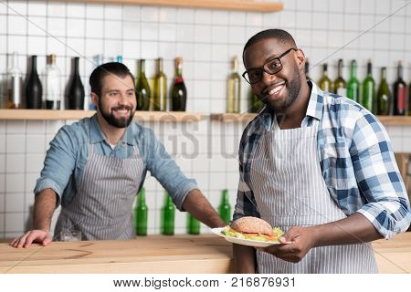 Working together. Positive young cheerful waiter holding a plate with a tasty fresh sandwich while his friendly colleague standing at the bar counter and looking at him