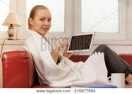 Young woman lying on sofa under warm blanket relaxing using tablet reading news and checking social media having fun during leisure time. Modern relaxation.