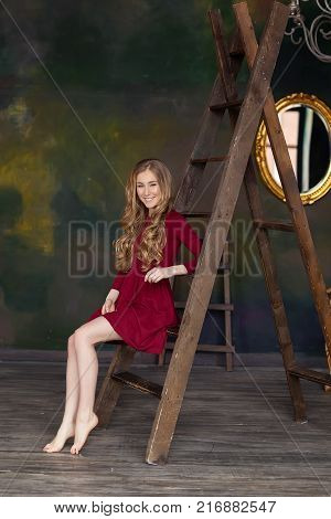 Beautiful young woman sitting on step ladder. Looking at camera. Beautiful face. Long blonde hair. Long legs. Short dress wearing.