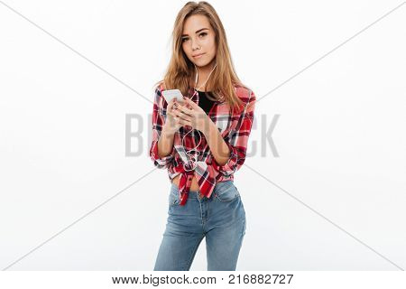 Portrait of a lovely sweet girl in plaid shirt holding mobile phone and listening to music with earphones isolated over white background