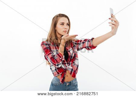 Portrait of a pretty lovely girl in plaid shirt taking a selfie and blowing air kiss isolated over white background