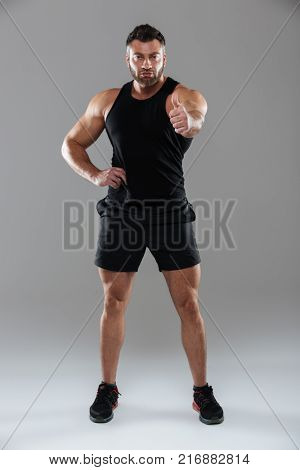 Full length portrait of a confident strong male bodybuilder standing and showing thumbs up gesture isolated over gray background