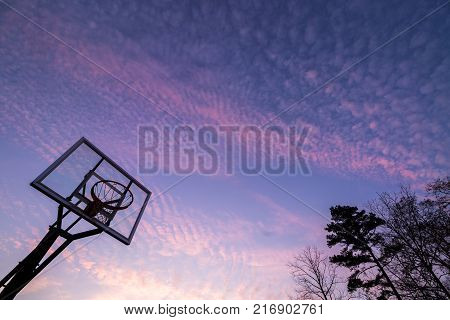 Silhouette of outdoor basketball goal with clear backboard and sunset in the background visible through backboard