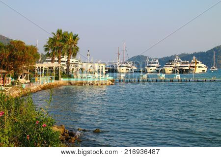 Photo taken in Turkey in Gocek. In the photo there is a cafe located on the seashore and pier. On the background of the cafe you can see the marina with yachts.