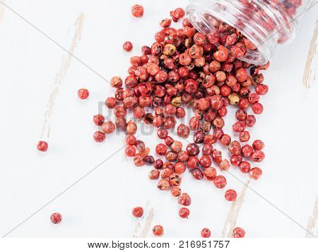 Dried pink pepper berries poured from glass jar on white wooden cutting board. Close up view of pink peppercorn poured on vintage wooden table. Copy space. Top view or flat-lay.