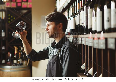 Bartender looks at red wine in shiny glass in cellar with large shelves full of exquisite delicious alcohol drinks and wooden barrel with bottle of champagne on top.