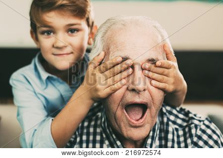 Portrait of little Caucasian boy making surprise covering eyes of senior grandfather at home