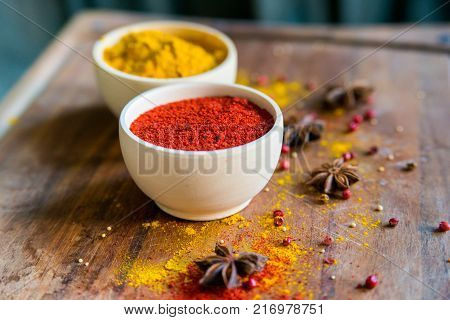 Close up two bowls filled with red paprika and yellow turmeric powder on dark wooden table covered with anise
