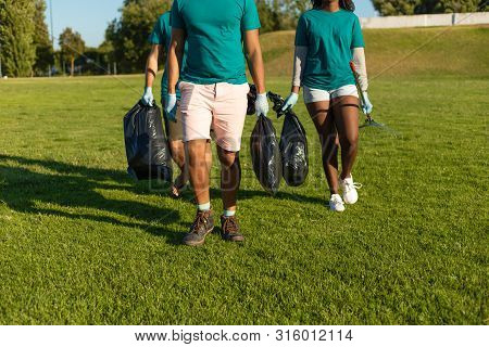 Cleaners Working Removing Rubbish From City Lawn. Young Woman And Men Walking On Grass And Carrying 
