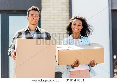 Cople Standing Near House, Looking At Camera And Holding Boxes