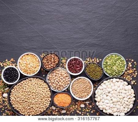 Legumes, lentils, chickpeas and beans assortment in different bowls on black stone background, top view with copy space