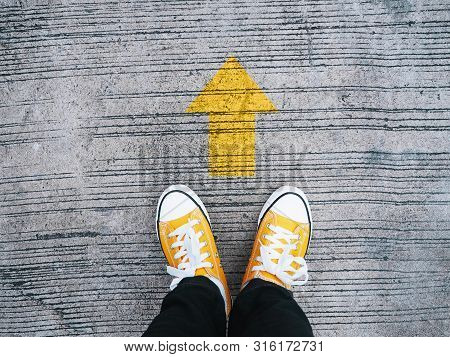 Selfie Feet Wearing Yellow Sneakers In Front Of Arrow On Concrete Road. Start Moving Forward Concept