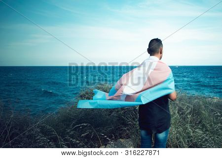 a young caucasian person, seen from behind, draping a transgender pride flag over his or her shoulders, facing the ocean