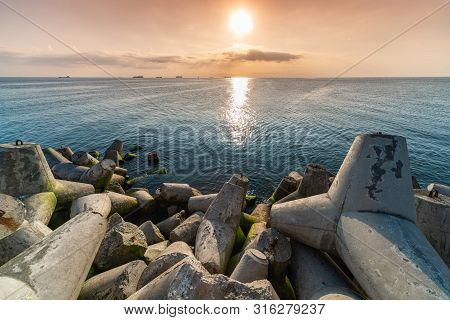 Beautiful Sunset Seascape. Breakwaters Tetrapods Ashore Of Pier. Cargo Ships On The Horizon. Travel 