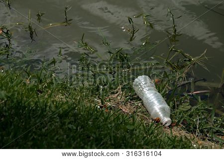 Plastic Waste Is Washed Up On The Lake Shore. A Plastic Bottle And Plastic Fork Rest Among The Grass