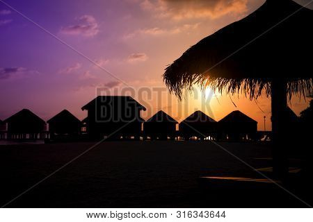 Colorful Sunset In Maldives With A Silhouette Of Parasol And Bungalows.