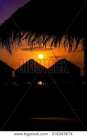 Colorful Sunset In Maldives With A Silhouette Of Parasol And Bungalows.