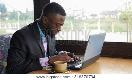Afro American Businessman Is Typing A Message On Laptop Sitting In Summer Cafe Near Window. Black Ma