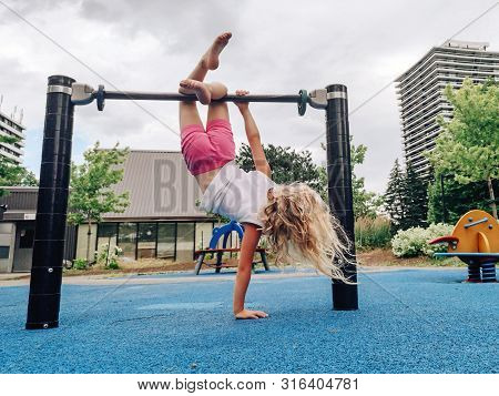 Cute Adorable Little Preschool Blonde Caucasian Girl Hanging Upside Down On Pullup Bar On Playground