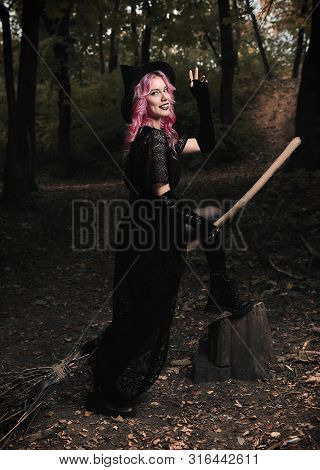 Halloween Theme: Modern Smiling Young Witch In Black Dress And Hat With Broom In A Dark Forest