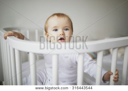 Adorable Baby Girl In Co-sleeper Crib Attached To Parents Bed. Little Kid Sitting In Cot