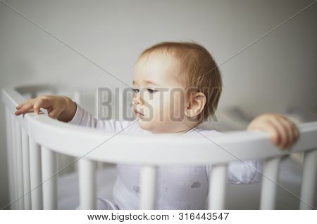 Adorable Baby Girl In Co-sleeper Crib Attached To Parents Bed. Little Kid Sitting In Cot