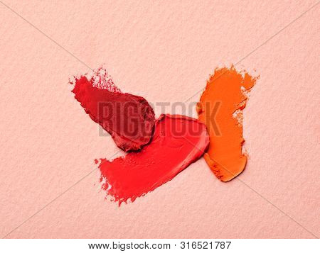 Various Color Lipstick Smears On Pale Pink Textured Background. Orange, Pink And Red Makeup Palette.