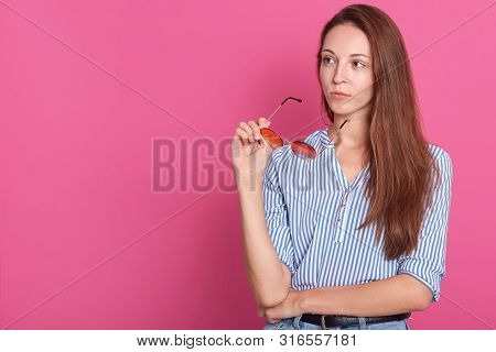 Studio Shot Of Young Woman Dessed Elegant Shirt, Holding Her Glasses In Hand, Looking Aside Isolated