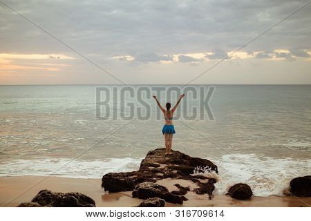 Excited Young Woman Standing On The Rock, Raising Arms At The Beach In Front Of The Ocean. View From
