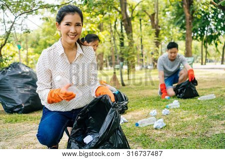 Young Asian Woman And Group Of Volunteers Wearing Orange Gloves And Collecting Garbage In Trash Bin 
