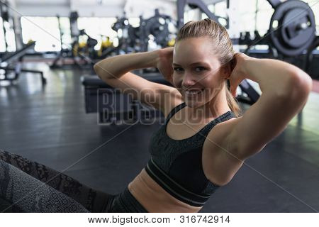 Side view of Happy Caucasian woman looking at camera while doing crunches in fitness center. Bright modern gym with fit healthy people working out and training