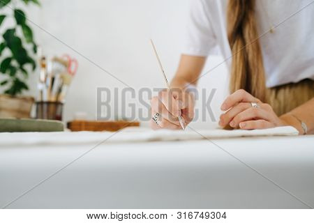 Young Woman Artist With Unfixed Braid Is Painting On Sheet, Behind The Table At Home. Cropped, No He