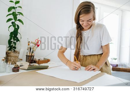 Young Smiling Focused Woman Artist With Unfixed Braid Is Drawing On Sheet, Behind The Table At Home.